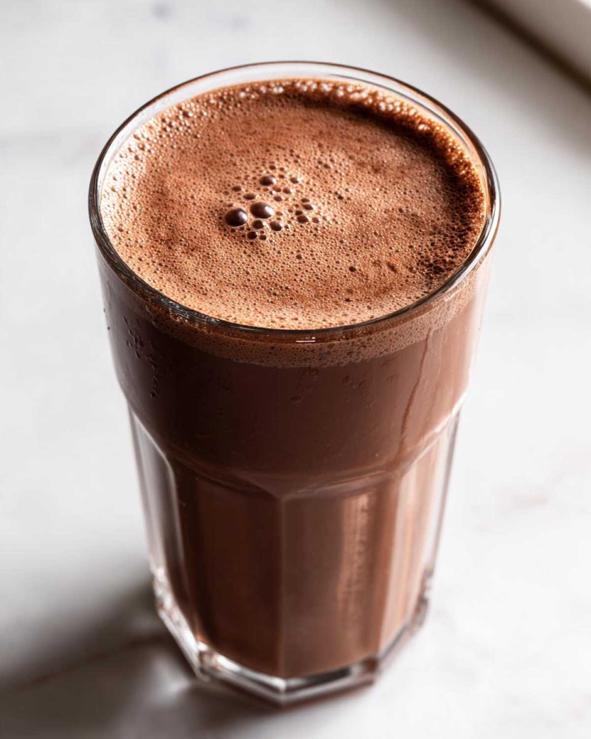 Overhead view of a tall glass filled with rich, dark chocolate milk topped with a layer of fine foam.