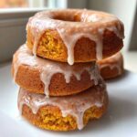 Three stacked, moist baked pumpkin donuts covered in a thick, dripping vanilla glaze on a white plate.