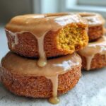A stack of moist baked pumpkin donuts topped with a thick, dripping caramel glaze, one donut has a bite taken out.