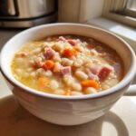A close-up of a white bowl filled with hearty bean soup featuring white beans, diced ham, and carrots.