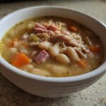 Close-up of a white bowl filled with hot navy bean soup, featuring white beans, shredded chicken, diced ham, and carrots.