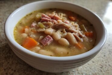 Close-up of a white bowl filled with hot navy bean soup, featuring white beans, shredded chicken, diced ham, and carrots.