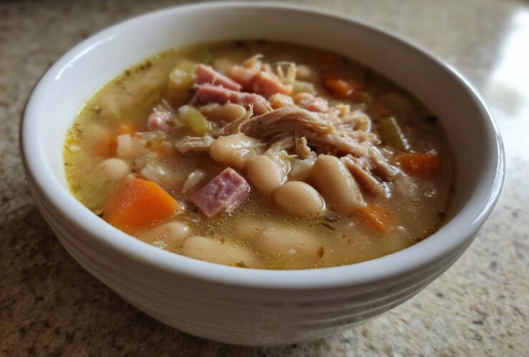 Close-up of a white bowl filled with hot navy bean soup, featuring white beans, shredded chicken, diced ham, and carrots.