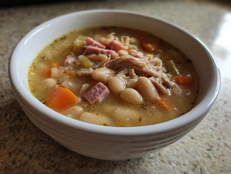 Close-up of a white bowl filled with hot navy bean soup, featuring white beans, shredded chicken, diced ham, and carrots.