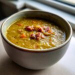 Close-up of a thick, green pea soup topped with diced ham and carrots in a light ceramic bowl.