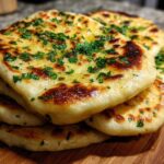 A stack of golden brown, fluffy homemade naan bread brushed with butter and topped with fresh green herbs.