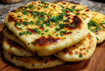 A stack of golden brown, fluffy homemade naan bread brushed with butter and topped with fresh green herbs.