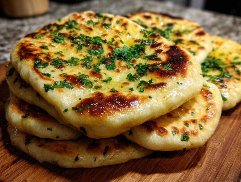 A stack of golden brown, fluffy homemade naan bread brushed with butter and topped with fresh green herbs.