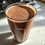 A tall glass filled with freshly made chocolate milk, showing a layer of small foam bubbles on top.