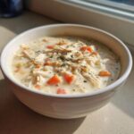 A white bowl filled with creamy homemade cream of chicken soup, featuring shredded chicken and diced carrots, garnished with herbs.