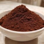 Close-up of a generous mound of rich, dark brown homemade hot cocoa mix powder overflowing a small white bowl.