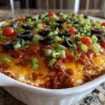 Close-up of a bubbling hot layer dip in a white baking dish, topped with melted cheese, black olives, tomatoes, and green onions.