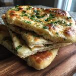 Close-up of stacked, golden-brown naan bread slices brushed with butter and topped with fresh green herbs.