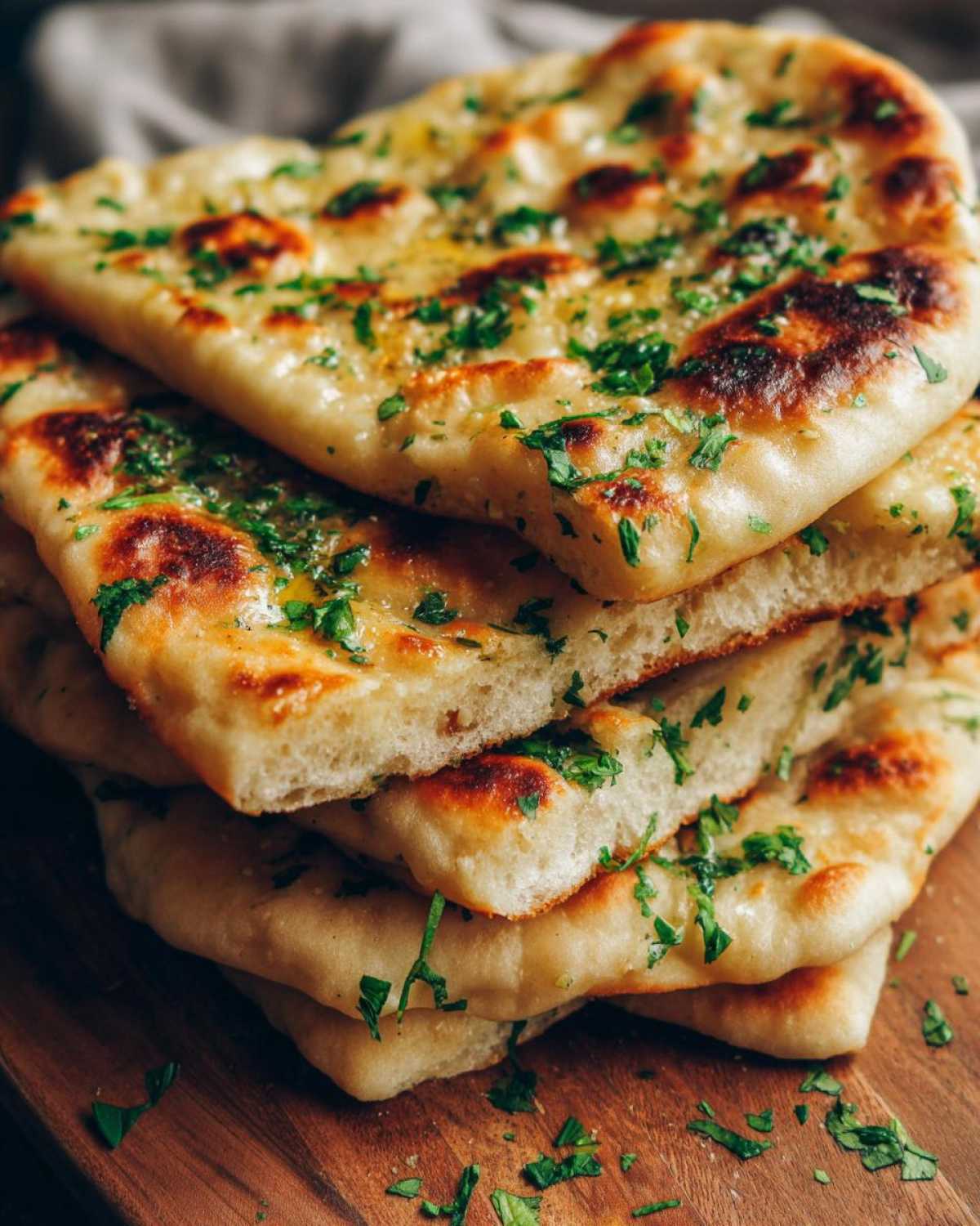 A close-up stack of fluffy, golden-brown naan bread brushed with butter and topped with fresh chopped cilantro.