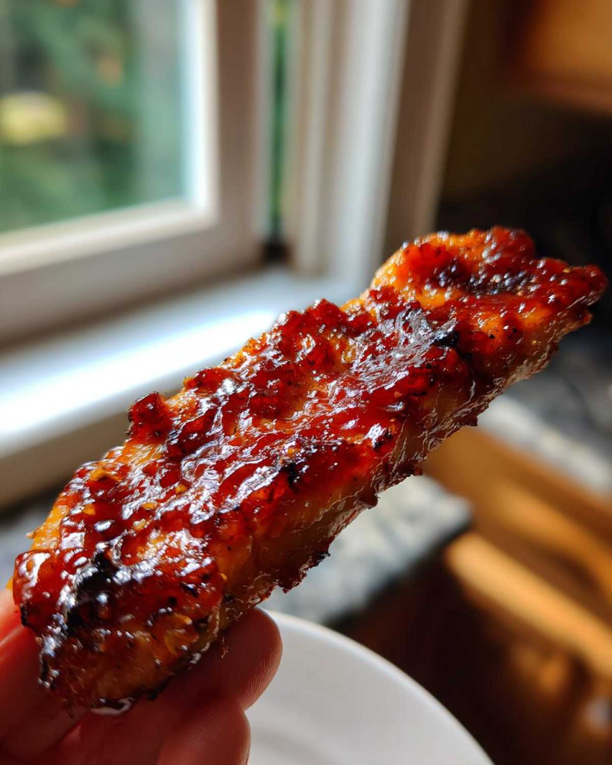 Close-up of a piece of glazed meat heavily coated in shiny, thick homemade bbq sauce, held up near a window.