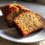 A close-up of a moist slice of homemade banana bread next to the rest of the loaf on a white plate.