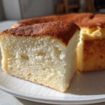 Close-up of a slice of moist, fluffy condensed milk cake with a golden brown top, sitting on a white plate.