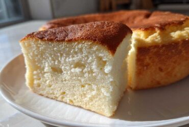 Close-up of a slice of moist, fluffy condensed milk cake with a golden brown top, sitting on a white plate.