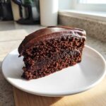 A close-up of a rich, moist slice of homemade chocolate cake topped with dark chocolate frosting on a white plate.