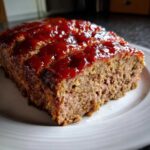 A thick slice of moist meatloaf topped with a shiny, red ketchup glaze, served on a white plate.