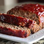 Close-up of a moist meatloaf, sliced and covered in a shiny, thick red glaze.