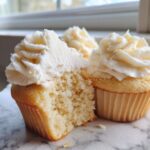 Close-up of a vanilla cupcake cut in half showing its moist crumb, topped with white vanilla frosting.
