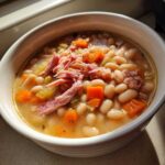 A close-up of a white bowl filled with hearty navy bean soup, featuring white beans, carrots, celery, and topped with shredded ham.