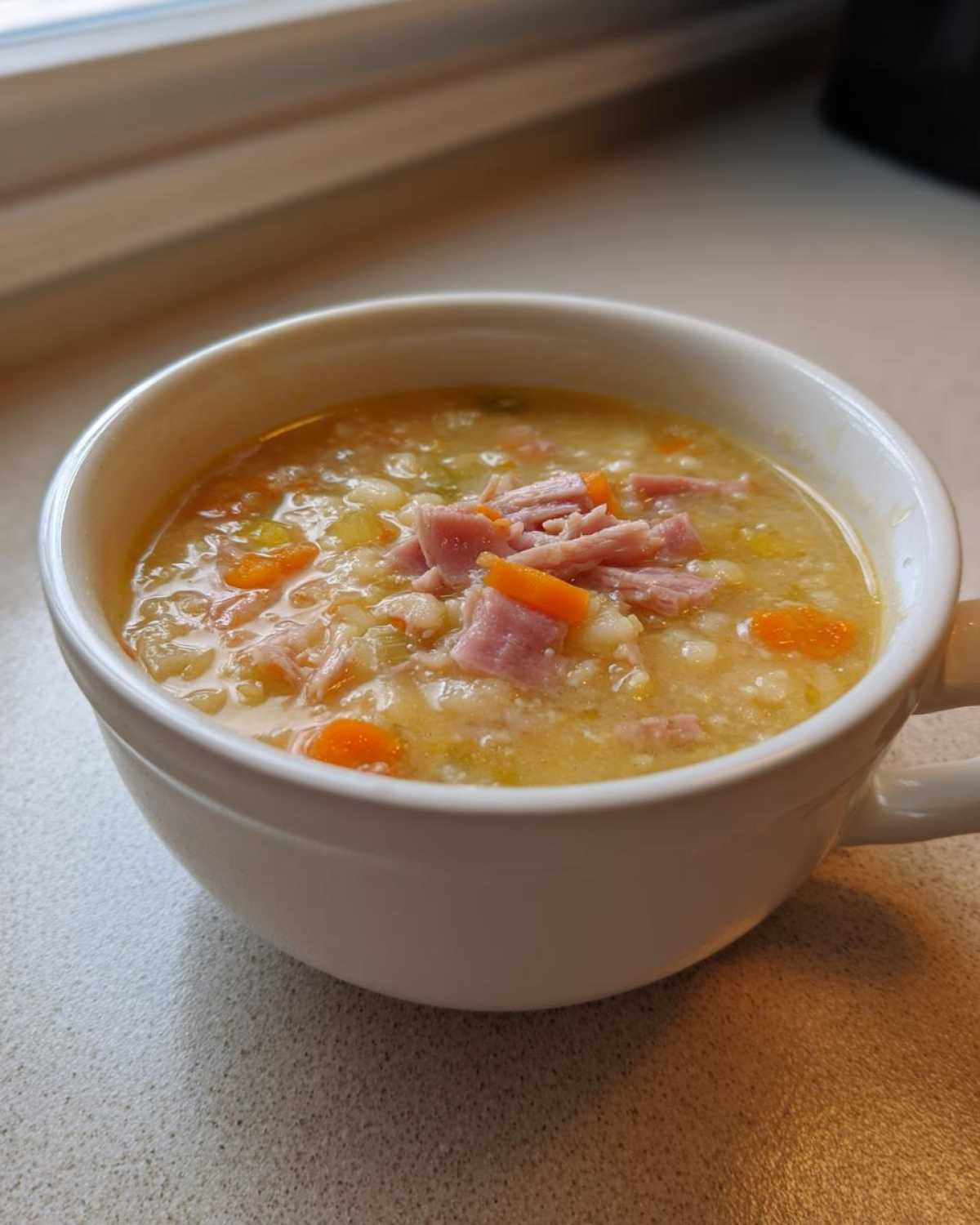 Close-up of a white mug filled with thick navy bean soup, topped with shredded ham and diced carrots.