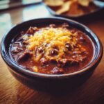 A close-up of a dark bowl filled with hearty cowboy soup, topped generously with shredded cheddar cheese.