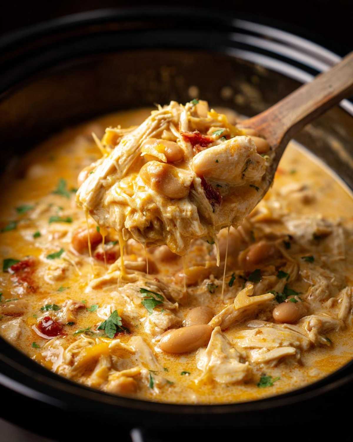 A wooden spoon lifts a generous scoop of creamy, shredded cream cheese chicken chili with white beans from a dark slow cooker.