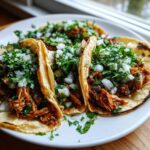 Close-up of three soft corn tortillas filled with shredded chicken tacos, topped generously with diced white onion and fresh cilantro.