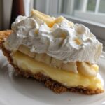 A close-up slice of banana cream pie featuring a graham cracker crust, thick vanilla custard, banana slices, and a generous topping of whipped cream.