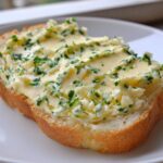 A thick slice of bread generously topped with creamy, bright yellow garlic butter mixed with fresh green herbs.