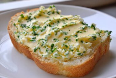 A thick slice of bread generously topped with creamy, bright yellow garlic butter mixed with fresh green herbs.