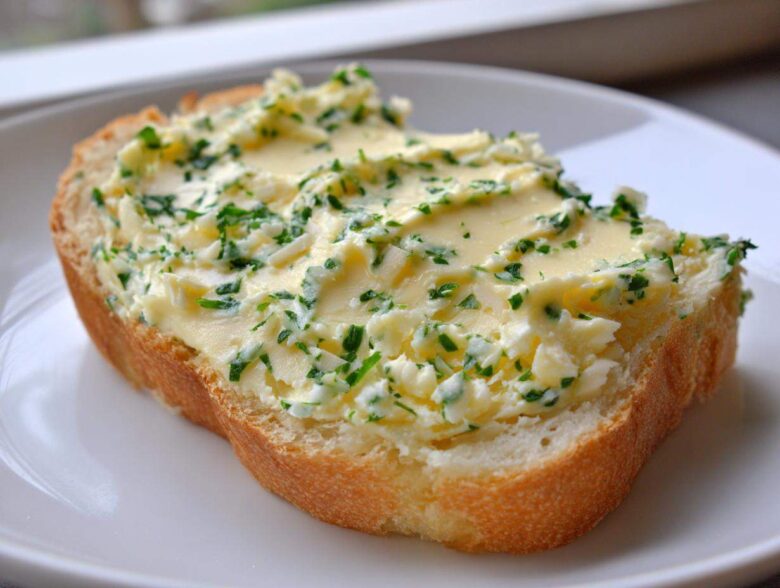 A thick slice of bread generously topped with creamy, bright yellow garlic butter mixed with fresh green herbs.