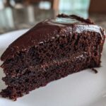 Close-up of a moist slice of chocolate fudge cake with thick, glossy dark chocolate frosting on a white plate.