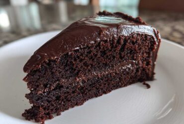 Close-up of a moist slice of chocolate fudge cake with thick, glossy dark chocolate frosting on a white plate.