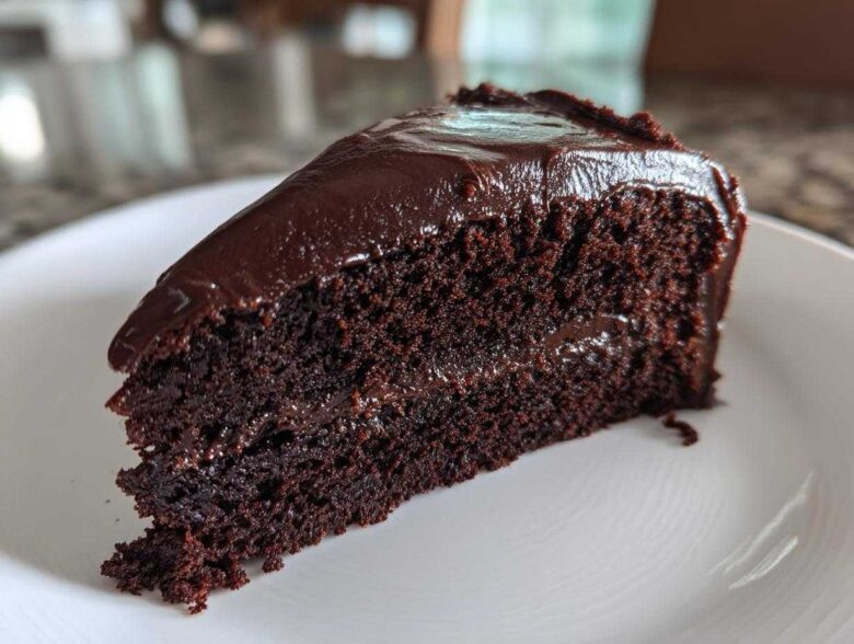 Close-up of a moist slice of chocolate fudge cake with thick, glossy dark chocolate frosting on a white plate.