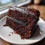 Close-up of a moist slice of chocolate fudge cake topped with thick, glossy chocolate frosting on a white plate.