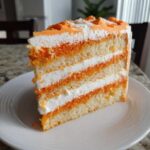 A tall slice of Creamsicle Cake showing four layers of white cake alternating with orange-tinted crumbs and white frosting.