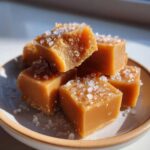 A stack of soft, chewy, square caramels topped generously with flaky sea salt, sitting on a small ceramic plate.