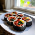 Six freshly cut pieces of spicy tuna kimbap arranged on a white plate near a sunny window.