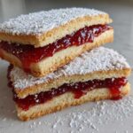 Two stacked, rectangular linzer cookies filled with bright red raspberry jam and dusted heavily with powdered sugar.