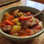 Close-up of a bowl of rich vegetable beef soup, featuring chunks of beef, carrots, potatoes, and celery.