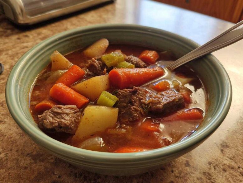 Close-up of a bowl of rich vegetable beef soup, featuring chunks of beef, carrots, potatoes, and celery.