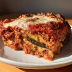 A close-up, sunlit slice of zucchini lasagna showing layers of meat sauce, cheese, and a visible zucchini noodle.