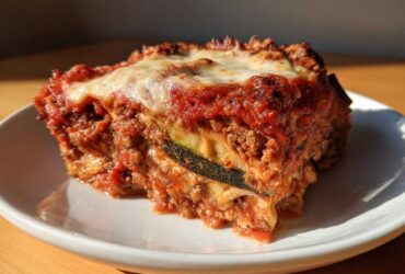 A close-up, sunlit slice of zucchini lasagna showing layers of meat sauce, cheese, and a visible zucchini noodle.