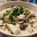 A close-up of a white bowl filled with creamy coconut soup featuring chunks of chicken, sliced mushrooms, and fresh cilantro.