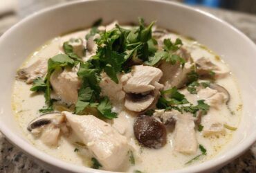 A close-up of a white bowl filled with creamy coconut soup featuring chunks of chicken, sliced mushrooms, and fresh cilantro.