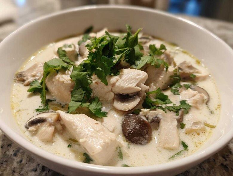 A close-up of a white bowl filled with creamy coconut soup featuring chunks of chicken, sliced mushrooms, and fresh cilantro.